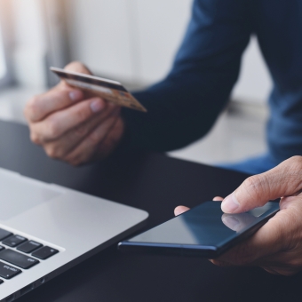 Ein Mann sitzt vor seinem Notebook und hält in der linken Hand sein Smartphone, in der rechten Hand eine Kreditkarte