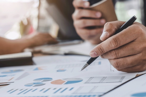 Ein Mann hält einen Kugelschreiber und betrachtet Unterlagen, eine Person im Hintergrund bedient ein Notebook