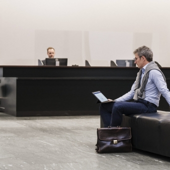 Ein Mann mit Brille sitzt auf einem Sessel in einer Bankfiliale und schaut auf seinem Tablet.