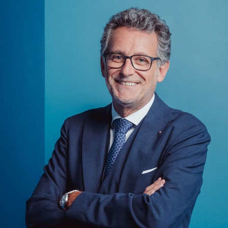 Ein Mann mit Brille, einem dunkelblauen Anzug mit blau gemusterter Krawatte sowie einem weißen Hemd lächelt